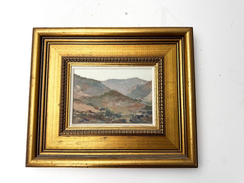May include: A framed landscape painting featuring a mountain range in muted tones of brown, green, and gray. The artwork is encased in a gold-colored wooden frame with a detailed inner border, creating a classic aesthetic. The painting is a small size.