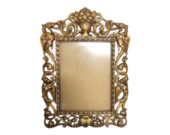 May include: Ornate gold-colored picture frame with a rectangular opening. The frame features elaborate scrollwork, mythical bird-like figures, and a decorative urn at the top. The frame is designed with a vintage aesthetic.