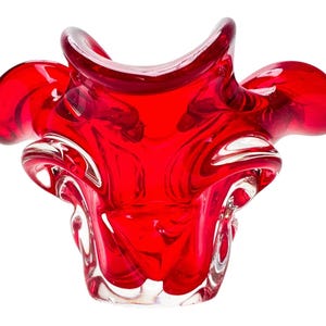 Puede incluir: Un cuenco de cristal rojo vibrante con una forma orgánica compleja, múltiples bordes ondulados y plegados, y una base de cristal transparente. El material translúcido capta la luz, resaltando el color carmesí intenso y la forma fluida.