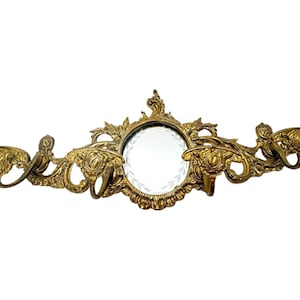 May include: Ornate gold-colored wall mirror with a round, clear glass mirror in the center. The mirror is surrounded by detailed, swirling floral and scrollwork designs. Two curved hooks are on either side of the mirror.