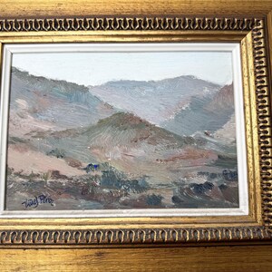 May include: Oil painting of a mountain landscape in muted tones of blue, green, and brown. The artwork is framed in a gold-colored, ornate frame with a white inner border. The artist's signature is in the lower left corner.