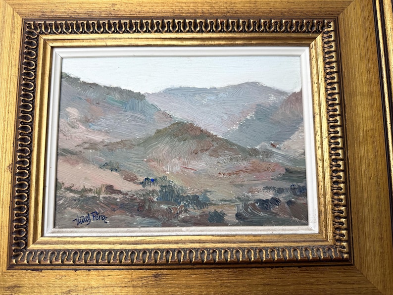 May include: Oil painting of a mountain landscape in muted tones of blue, green, and brown. The artwork is framed in a gold-colored, ornate frame with a white inner border. The artist's signature is in the lower left corner.