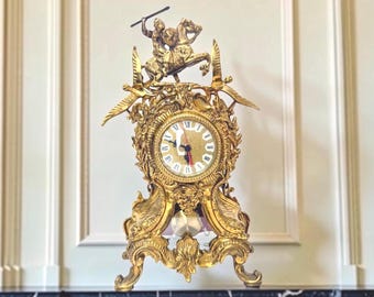 May include: A gold-toned mantel clock featuring an ornate frame with intricate scrollwork and foliage motifs. The top displays a figure of a mounted warrior holding a spear, flanked by two birds. The clock face has Roman numerals and black hands.