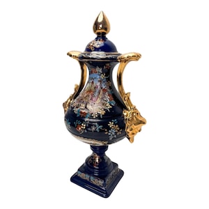 Peut inclure: Une urne en céramique bleu profond avec un couvercle amovible surmonté d'un fleuron doré. Le récipient possède deux anses dorées ornées en forme de têtes de lion, et le corps est décoré de motifs floraux colorés et d'une scène paysagère centrale.