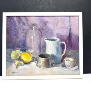 May include: Oil painting in a white frame depicting a still life. A clear bottle, a white pitcher, a small silver cup, and a teacup are arranged on a table with a lemon and leaves. The background is purple.