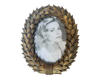 May include: Oval gold-toned picture frame with a leaf design. The frame surrounds a black and white photo. The frame is approximately 5" x 7" and has a rope-like detail at the bottom.