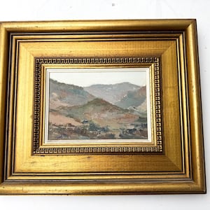 May include: A framed landscape painting featuring a mountain range in muted tones of brown, green, and gray. The painting is set within a gold-colored, ornate frame, adding a touch of elegance to the artwork. The frame measures approximately 12 x 10 inches.