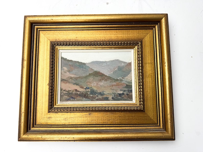 May include: A framed landscape painting featuring a mountain range in muted tones of brown, green, and gray. The painting is set within a gold-colored, ornate frame, adding a touch of elegance to the artwork. The frame measures approximately 12 x 10 inches.