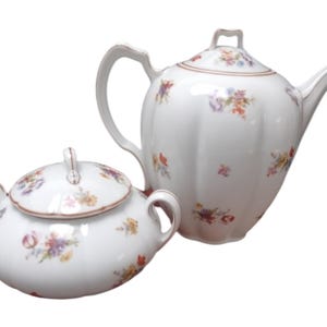 Op de afbeelding: Witte porseleinen theeset met bloemaccenten. De set bevat een theepot met een gebogen tuit en handvat, en een suikerpot met een deksel en twee handvatten. Het bloemmotief toont roze, gele en paarse bloemen.
