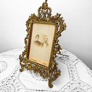 May include: Ornate antique brass picture frame with a sepia-toned photograph of a couple. The frame has intricate floral and scrollwork details, supported by a stand. The photograph is rectangular, with a dark border. The frame is displayed on a white, lace-like surface.