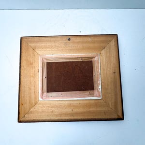 May include: Back of a rectangular wooden picture frame. The frame is made of light brown wood with a darker brown trim. The back of the frame has a brown backing and a small metal hook for hanging.