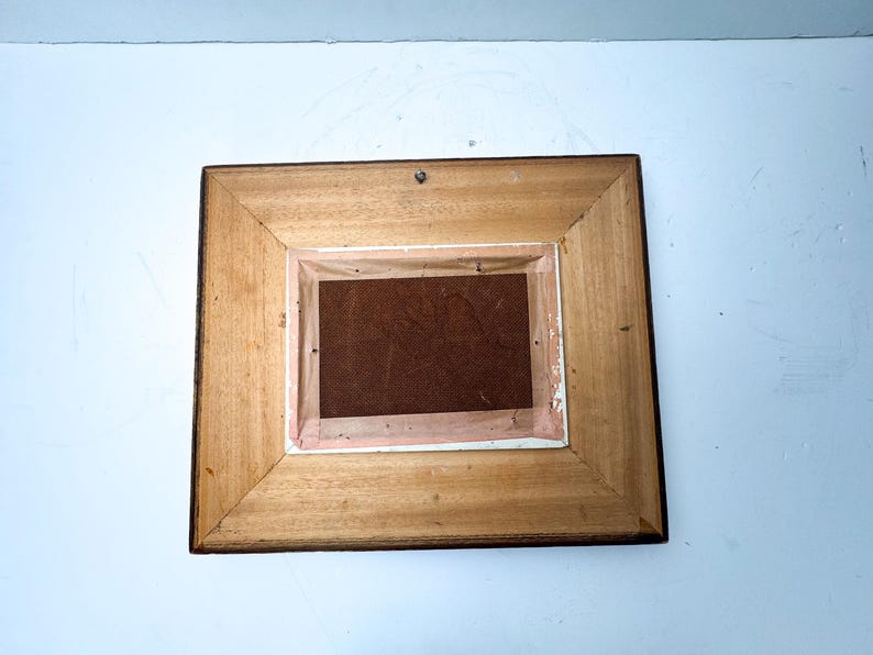 May include: Back of a rectangular wooden picture frame. The frame is made of light brown wood with a darker brown trim. The back of the frame has a brown backing and a small metal hook for hanging.