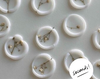 Peut inclure: Gros plan sur des sceaux de cire blancs avec des accents floraux séchés. Chaque sceau rond contient une délicate brindille de fleurs séchées. Le texte "Seconds!" est écrit sur un cercle blanc en bas à droite.