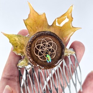 Op de afbeelding: Een unieke haar kam met een bewaard gebleven esdoornblad in geel- en groentinten. De kam heeft een centraal bruin element met een gouden geometrisch ontwerp en een kleine iriserende edelsteen. Zilveren metalen tanden maken het ontwerp compleet.