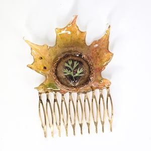 Op de afbeelding: Een haar kam met een esdoornblad ontwerp. Het blad is een mix van geel, groen en bruin, met een cirkelvormige inzet met een kleine plant. De tanden van de kam zijn goudkleurig.