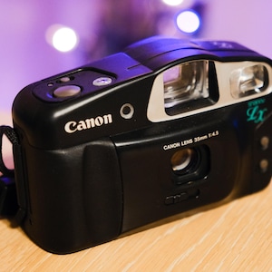 May include: A black Canon Snappy LX 35mm film camera featuring a Canon Lens 35mm 1:4.5. The camera body includes a built-in flash, a viewfinder, a shutter button on the top, and a black wrist strap attached to the side of the compact plastic housing.