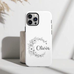 May include: A white smartphone case featuring a black line art floral wreath design arranged in a circular shape. The name Olivia is printed in a black script font inside the center of the floral wreath. The case has a cutout for a triple camera.