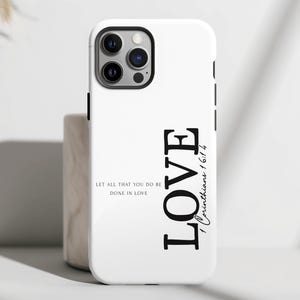 May include: White phone case with the word "LOVE" in large, bold, black letters. The case also features the text "Let all that you do be done in love" and "1 Corinthians 16:14". The phone case has a black camera module.