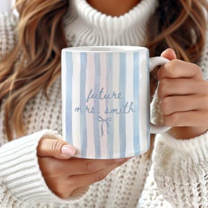 May include: White ceramic mug with blue and pink vertical stripes. The mug features the text "future mrs. smith" in light blue script, with a bow illustration below. The mug has a white handle.