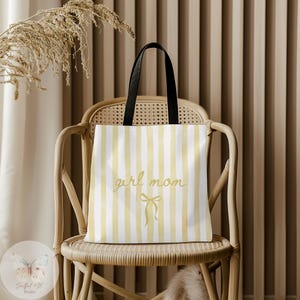 Puede incluir: Un bolso tote con rayas amarillas y blancas y un asa negra. Las palabras "girl mom" están escritas en cursiva, con un lazo debajo. El bolso está sobre una silla de mimbre.