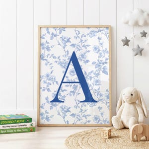 Puede incluir: Una lámina decorativa rectangular en un marco de madera clara que presenta una letra A mayúscula grande de color azul oscuro sólido centrada sobre un fondo blanco con un patrón floral azul claro repetitivo de flores delicadas, hojas y tallos finos.