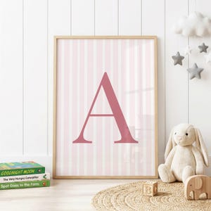 May include: A framed wall art print featuring a large, dark pink capital letter A centered on a background of vertical light pink and white stripes. The print is housed in a light-colored wooden frame, displaying a clean and simple design.