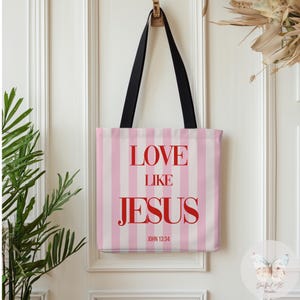 Puede incluir: Un bolso tote con un diseño de rayas rosas y blancas. El bolso presenta las palabras "LOVE LIKE JESUS" en rojo, con "JOHN 13:34" debajo. Tiene asas negras y está colgado de una puerta blanca.