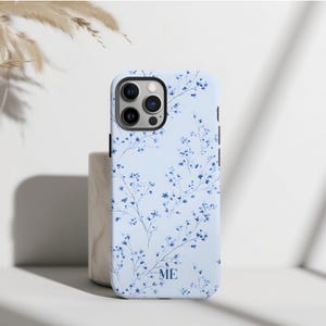 May include: A light blue phone case with a floral design. The case features a pattern of delicate blue flowers and stems. The initials "ME" are printed on the lower part of the case. The case is designed to fit a smartphone.