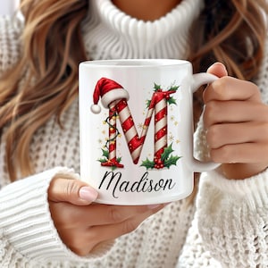 May include: A white ceramic mug featuring a large letter M designed to look like a red and white candy cane. The letter is decorated with a red Santa hat, green holly leaves, red berries, and small yellow stars. The name Madison is printed below.