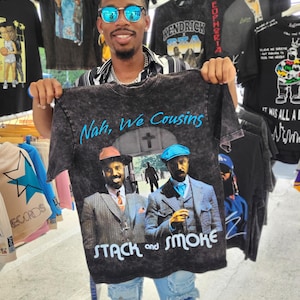 May include: A dark gray t-shirt with the text "Nah, We Cousins" and "STACK and SMOKE" in white. The shirt features a graphic of two men in suits. Other t-shirts with graphics are visible in the background.
