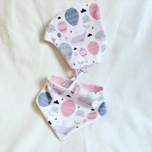 May include: A baby bib and face mask set. The items display a pattern of pink and grey hot air balloons, whales, and clouds on a white background. The bib is reversible with a pink interior.