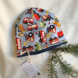 May include: A light blue beanie hat with a pattern of red fire trucks, yellow construction vehicles, and green ambulances. The hat has a dark blue trim and a tag attached. The hat is likely for a child.