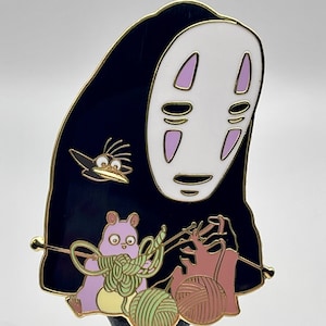 May include: A gold-tone enamel pin featuring No Face from the movie Spirited Away. No Face is wearing a black robe and holding yarn balls and knitting needles. A small purple cat and a black bird are also featured on the pin.