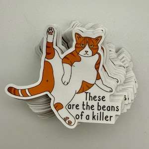 May include: A pile of stickers showing a cartoon cat in orange and white, with the words "These are the beans of a killer." The cat is in a playful pose, with one leg extended. The stickers have a white background.