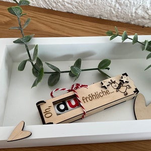 Könnte beinhalten: Eine Holzkiste mit einem roten und weißen Band darum gebunden. Die Kiste hat die Worte "Oh!" und "fröhliche..." darauf geschrieben. Es gibt eine kleine Rentier-Illustration auf der Kiste. Die Kiste steht auf einer weißen Oberfläche.