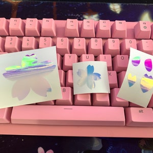 May include: Three sheets of white paper with holographic stickers of pink and blue flowers. The stickers are iridescent and have a rainbow effect.