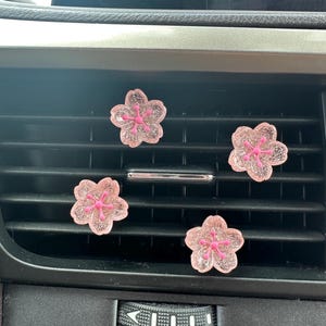 May include: Four flower-shaped car air vent clips featuring a translucent light pink glitter finish and a solid bright pink center stamen. These decorative accessories are attached to the horizontal slats of a black vehicle air conditioning vent.