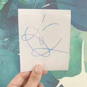 Op de afbeelding: Een witte sticker met een hartvormig ontwerp in blauw, roze en paars. Het hart is gemaakt van &eacute;&eacute;n doorlopende lijn.
