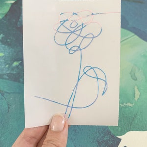 Op de afbeelding: Een sticker met een blauw en roze bloemetjesontwerp. De bloem is getekend in een eenvoudige, abstracte stijl.