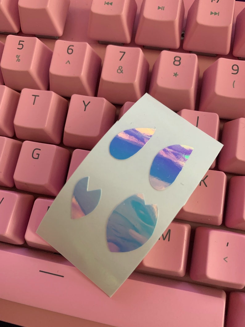 May include: A sheet of iridescent holographic stickers with a variety of shapes, including teardrop, heart, and leaf designs. The stickers are on a pink keyboard.