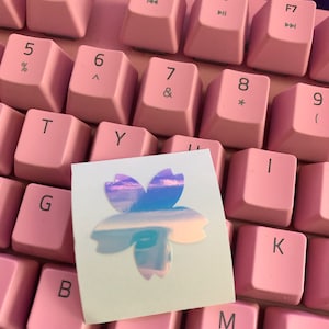 May include: A white sticker with a holographic pink and blue cherry blossom design on a pink keyboard.