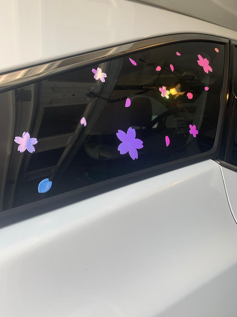 May include: A white car with a back window covered in pink and purple cherry blossom stickers. The stickers are arranged in a random pattern and are all different sizes.