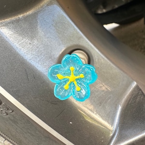 May include: A blue flower-shaped tire valve stem cap with a yellow center. The cap is made of plastic and has a sparkly finish.
