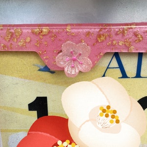 May include: A pink resin accent piece with gold flakes and a clear flower embellishment. The piece is set against a floral illustration featuring a red and white flower with the number 1.