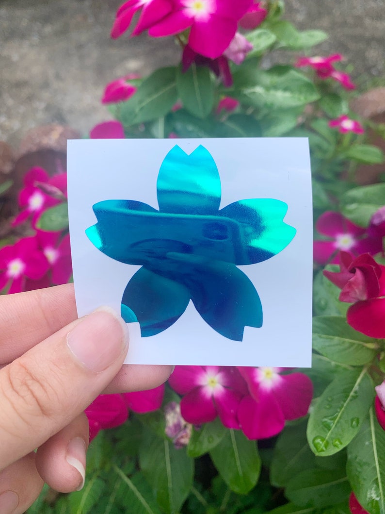 May include: A teal blue iridescent vinyl sticker of a five-petal flower, resembling a cherry blossom, on a white background.