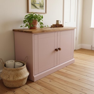 May include: A pale pink cabinet with two doors and wooden handles. It has a light wood top and stands on a wooden floor. A woven basket with rolled towels sits in front. A potted plant and books are on top.