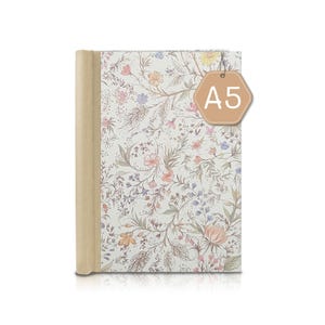 Peut inclure: Journal A5 floral avec un dos beige. La couverture présente un motif délicat de fleurs roses, jaunes et bleues avec des feuilles vertes sur fond blanc. Une étiquette beige avec le texte "A5" est suspendue en haut.