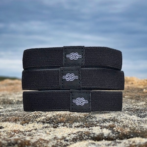 May include: Three black elastic hair ties with a white knot logo. The hair ties are stacked on top of each other on a rocky surface.