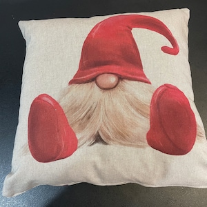 May include: A beige throw pillow featuring a red and white gnome design. The gnome is wearing a red hat and has a white beard.