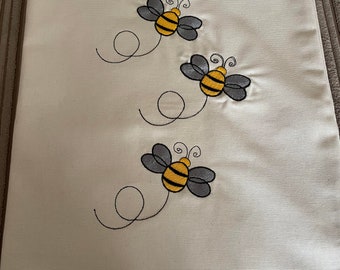 May include: A white canvas tote bag with three embroidered bees. The bees are yellow and black with gray wings. Each bee has a small swirl behind it.
